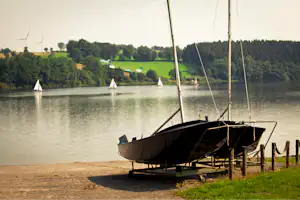 Ein Segelboot am Steg am See, im Hintergrund weitere Segelboote und grüne Ufer mit Bäumen.