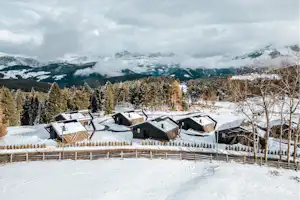 Winterliche Ferienhäuser auf einem Campingplatz mit verschneiten Wegen, Holzzaun und Blick auf verschneite Berge und Tannen.
