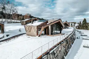 Mehrere Camping-Hütten mit Holzveranda stehen auf verschneitem Gelände neben einem Steinmauer und Zaun.
