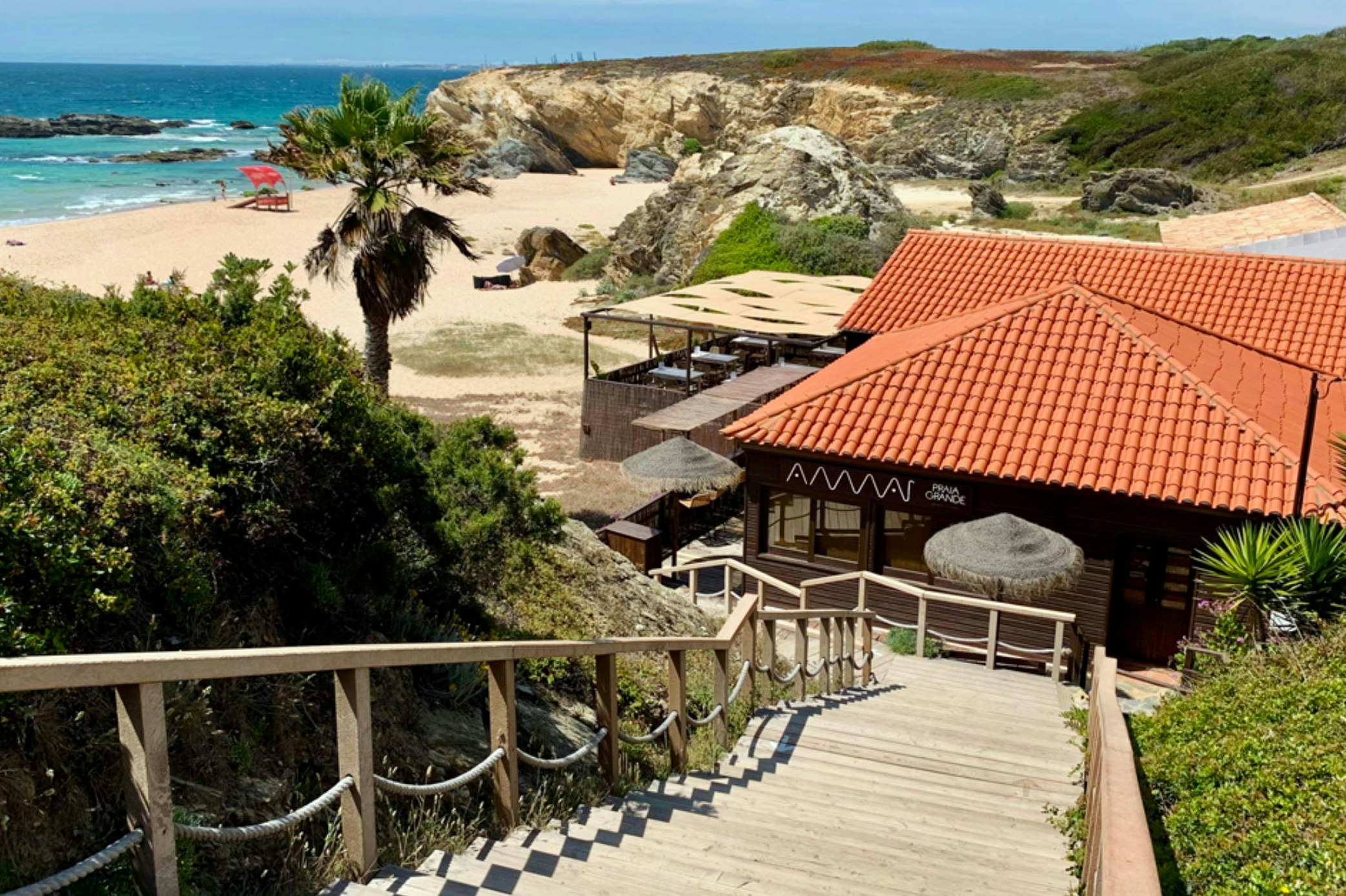 Terrasse mit Geländer und Sitzbereich am Campinghaus mit rotem Ziegeldach, Blick auf das Meer und den Strand in der Ferne.