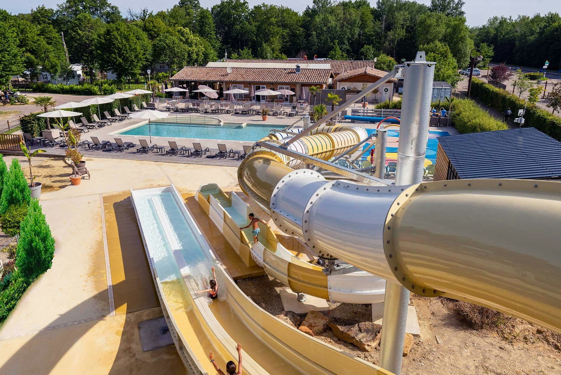 Blick auf den Camping-Pool mit Wasserrutsche, Liegen und Sonnenschirmen, im Hintergrund Bäume und Gebäude.