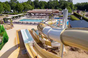 Blick auf den Camping-Pool mit Wasserrutsche, Liegen und Sonnenschirmen, im Hintergrund Bäume und Gebäude.