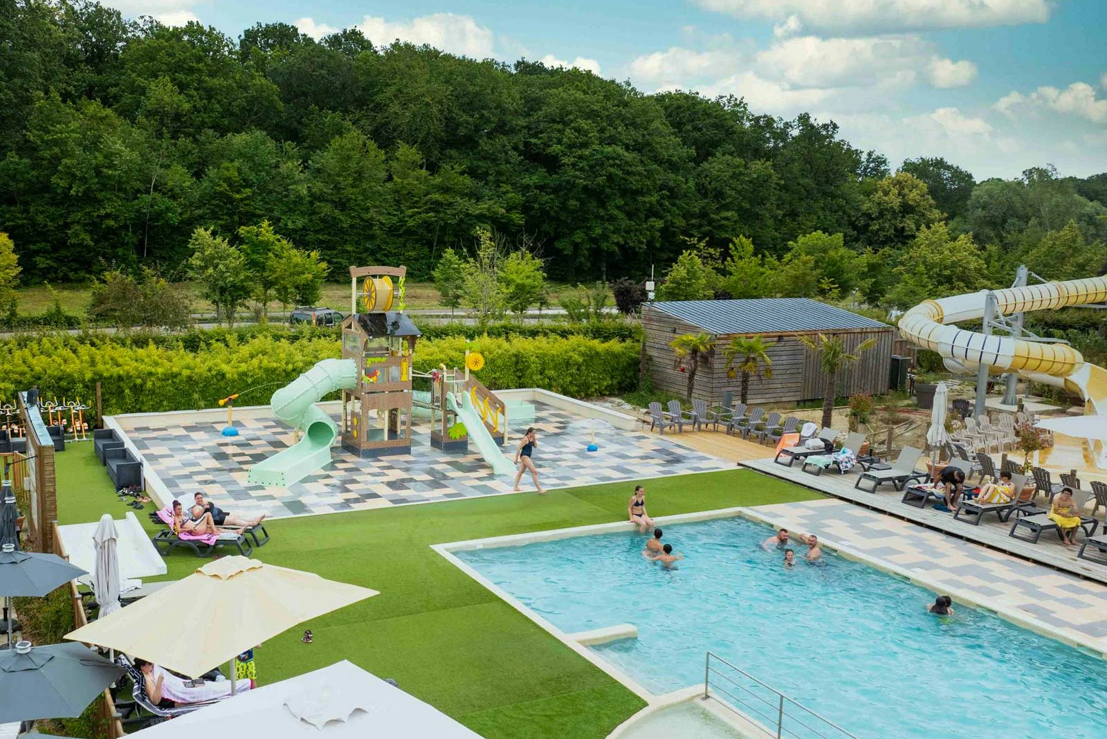 Großes Camping-Schwimmbecken mit Liegen, Sonnenschirmen und gepflasterter Terrasse, im Hintergrund Bäume und ein Steingebäude.