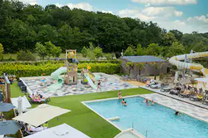 Großes Camping-Schwimmbecken mit Liegen, Sonnenschirmen und gepflasterter Terrasse, im Hintergrund Bäume und ein Steingebäude.