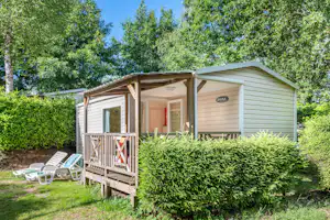 Stellplatz mit Wohnwagen und Holzveranda, davor Gartenhecke und zwei Liegestühle im Grünen am Campingplatz.
