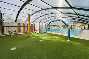 Überdachter Poolbereich mit grünem Kunstrasen, Spielgeräten und blauen Wasserrutschen, dahinter ein Zaun und Liegen.
