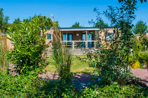 Moderner Bungalow im Campingbereich mit Terrasse, Glasfront und Blick ins Grüne, umgeben von Büschen und Rasen.