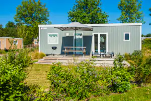 Bungalow mit Terrasse, Grill und Sitzbank unter Markise, umgeben von Hecken und Rasen im Grünen.