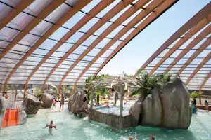 Schwimmbecken im Wasserpark unter einer Holzpergola mit Sonnensegeln, große Felsbrocken und Pflanzen im Hintergrund.