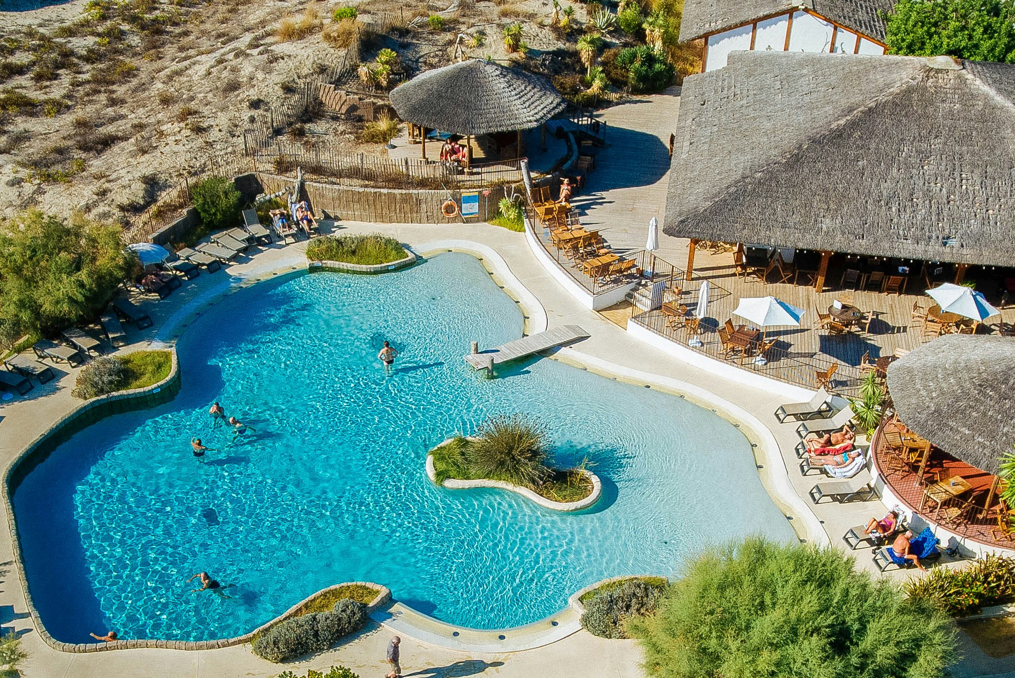 Großer Camping-Pool mit Sonnenschirmen, Liegen und Sitzbereichen, umgeben von Natursteinmauern und Palmen im Außenbereich.