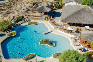 Großer Camping-Pool mit Sonnenschirmen, Liegen und Sitzbereichen, umgeben von Natursteinmauern und Palmen im Außenbereich.