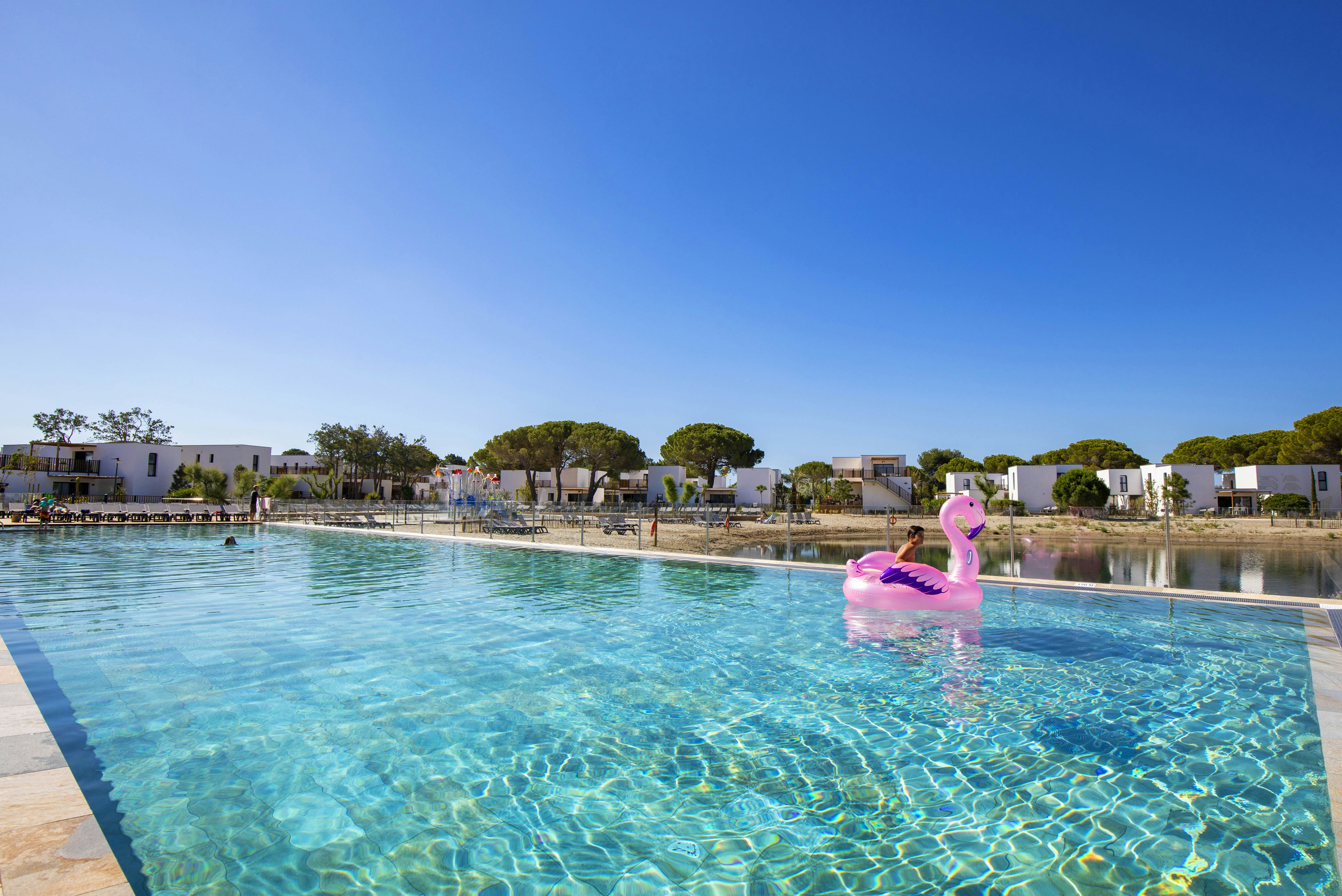Schwimmbecken mit klarem Wasser, rosa Schwan-Schwimmring und Blick auf Reihen weißer Ferienhäuser mit Terrassen.