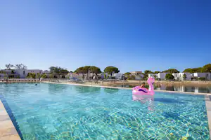 Schwimmbecken mit klarem Wasser, rosa Schwan-Schwimmring und Blick auf Reihen weißer Ferienhäuser mit Terrassen.