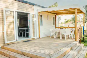 Holzbungalow mit Terrasse, Markise und weißen Gartenstühlen, Blick auf den Eingang und den Holzboden.