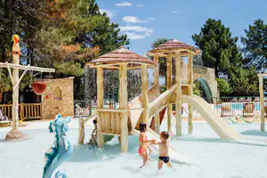 Wasserspielplatz auf dem Campingplatz mit Rutsche, gelbem Spielhaus und Wassersäulen vor Steinmauer und Bäumen.