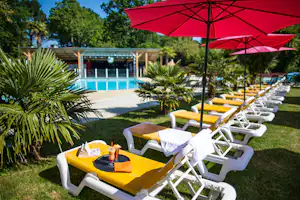 Gelbe Liegen und rote Sonnenschirme am Campingplatz, im Hintergrund ein Pool und ein Gebäude mit Terrasse.