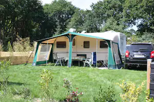 Wohnwagen mit grün-weißem Vorzelt, Campingstühlen und Tisch auf dem Rasen, daneben ein Auto und viel Grün.