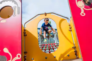 Ein Junge spielt in einem gelben Kinderfahrzeug mit roten Knöpfen und Metallgeländer, im Hintergrund ist ein Himmel zu sehen.