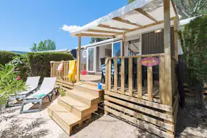 Holzbungalow mit Terrasse, Sonnensegeln, zwei Liegestühlen und Sichtschutzzaun im Garten, im Hintergrund Bäume und Sträucher.