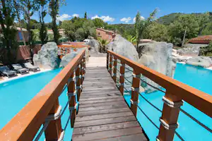 Holzsteg über dem türkisfarbenen Pool mit Geländer, Seilen und großen Felsen im Außenbereich, daneben Liegen.