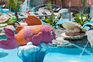 Buntes Wasserspiel mit Delfin- und Fischfiguren im Pool, im Hintergrund Liegen und Palmen am Campingplatz.