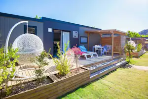 Moderner Campingbungalow mit Holzterrasse, Sonnenliegen und Gartenbeeten; im Vordergrund ein Whirlpool unter einer transparenten Kuppel.