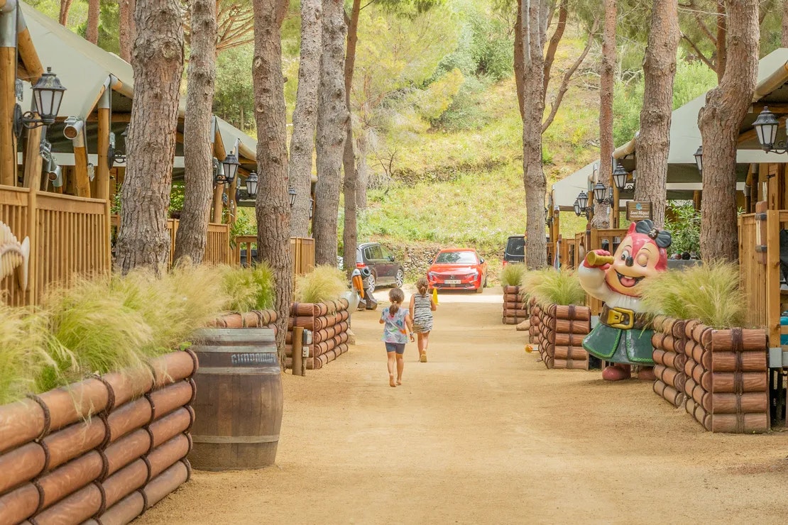 Weg im Campingplatz zwischen Baumstämmen, Holzgeländer und Sitzgelegenheiten, im Hintergrund ein Auto und ein Kind mit Rucksack.