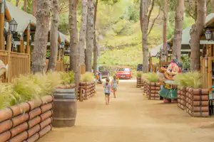 Weg im Campingplatz zwischen Baumstämmen, Holzgeländer und Sitzgelegenheiten, im Hintergrund ein Auto und ein Kind mit Rucksack.
