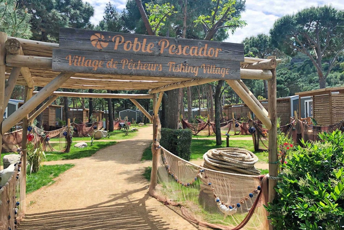 Eingangsschild „Poble Pescador“ am Campingweg, mit Holzgeländer, Hängematte und Bäumen im Hintergrund.