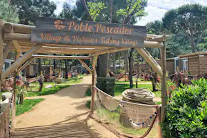 Eingangsschild „Poble Pescador“ am Campingweg, mit Holzgeländer, Hängematte und Bäumen im Hintergrund.