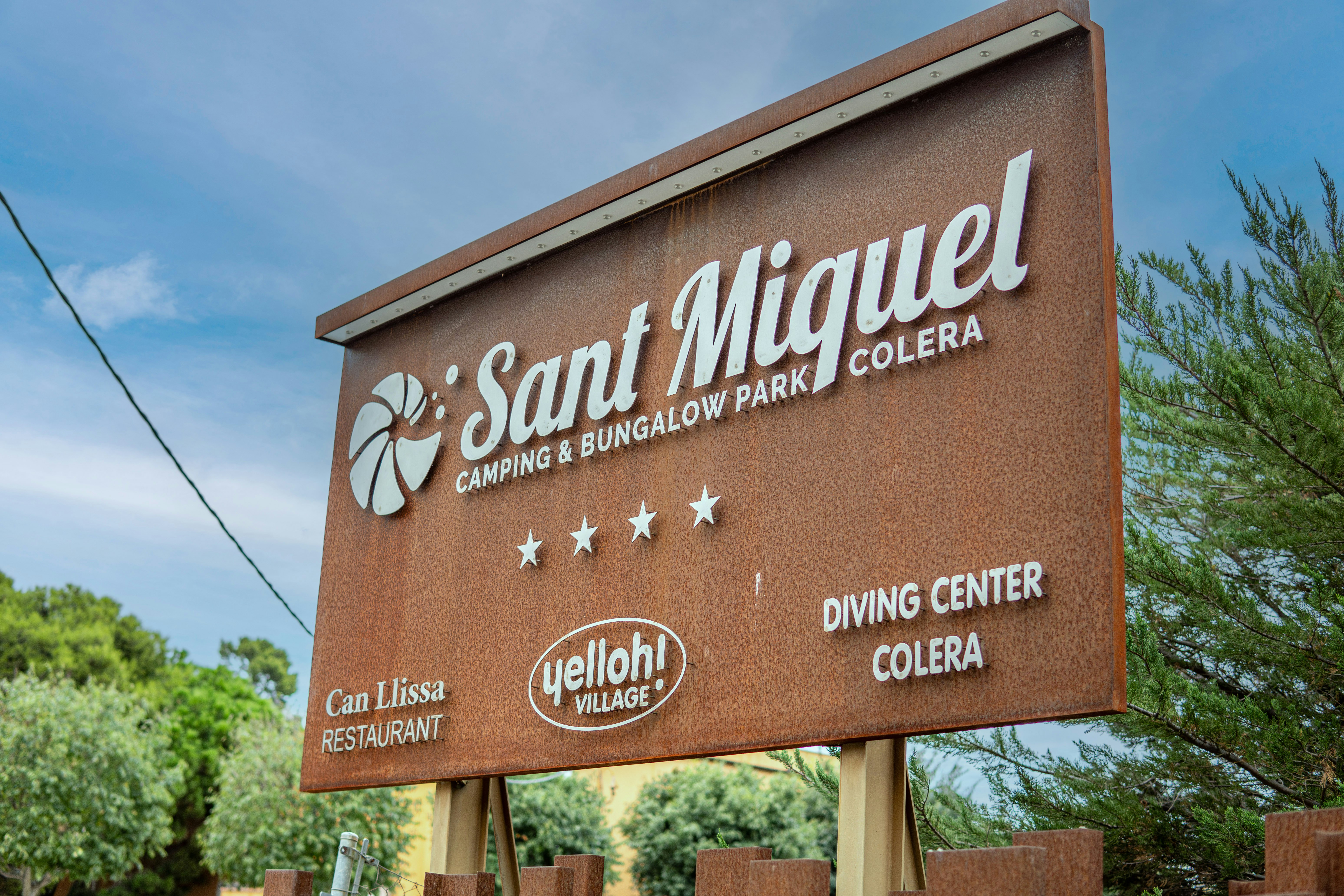 Schild am Campingplatz Sant Miquel mit Aufschrift Colera, Can Llissa Restaurant und Diving Center, umgeben von Bäumen und Himmel.