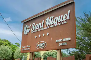 Schild am Campingplatz Sant Miquel mit Aufschrift Colera, Can Llissa Restaurant und Diving Center, umgeben von Bäumen und Himmel.