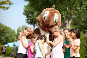 Kinder lachen und schauen auf ein großes Plüschtier mit grünem Auge und rotem Maul im Freien.