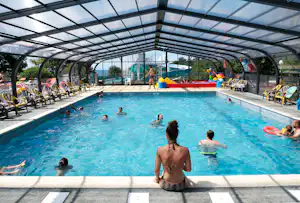 Überdachtes Camping-Schwimmbecken mit mehreren Liegen, Sonnenschirmen und badenden Gästen im Wasser.