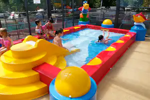 Kinder planschen in einem bunten Aufblas-Planschbecken auf dem Campingplatz, mit gelben Rutschen und Spielgeräten im Hintergrund.