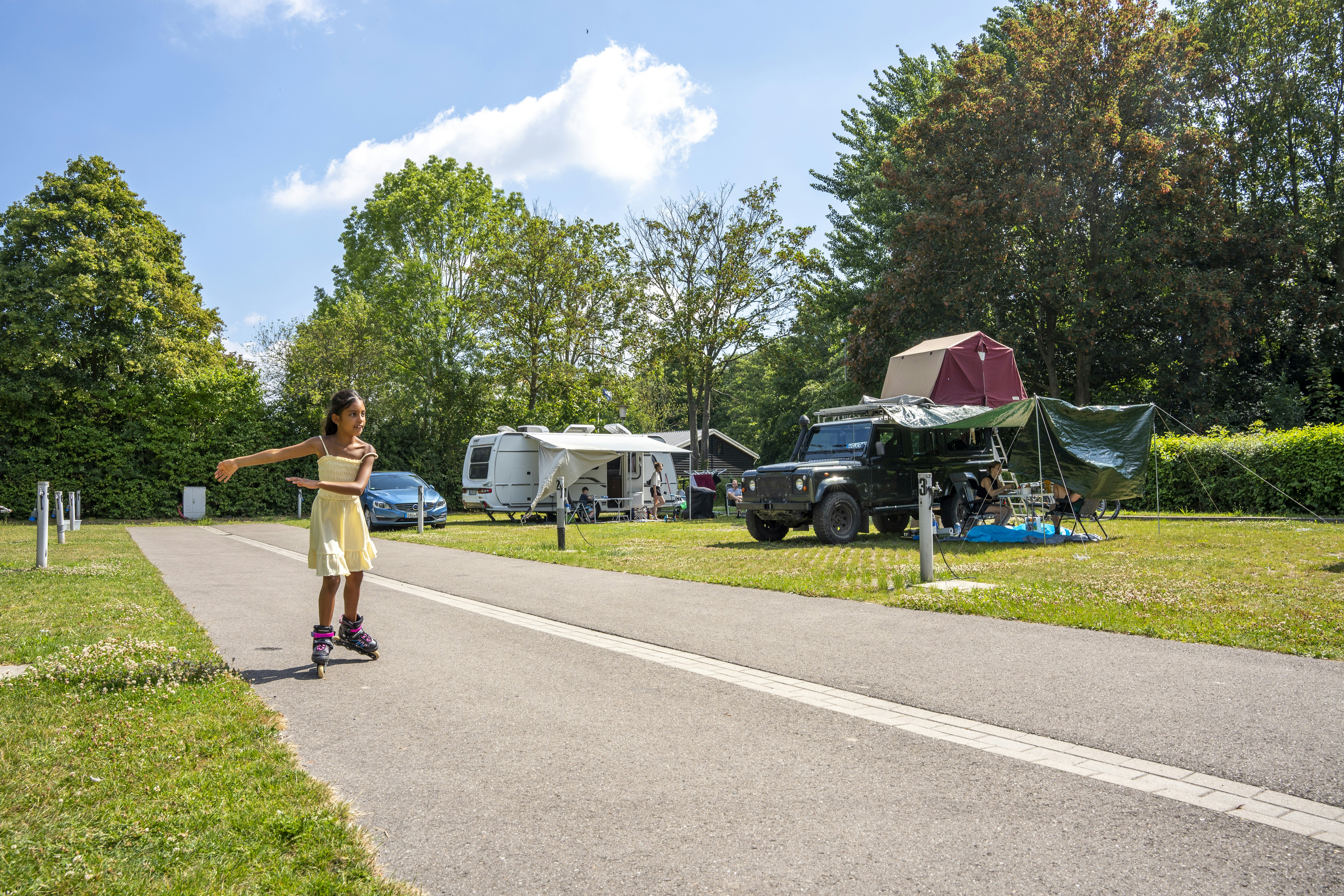 Ein Kind läuft auf dem Weg am Campingplatz, neben Wohnwagen, Auto und Zelt zwischen Bäumen und Rasen.