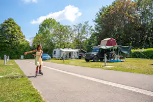 Ein Kind läuft auf dem Weg am Campingplatz, neben Wohnwagen, Auto und Zelt zwischen Bäumen und Rasen.