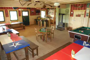 Spielraum im Campinghaus mit Billardtisch, roten und blauen Tischplatten, Stühlen und Holzdecke, im Hintergrund eine Küchenzeile.