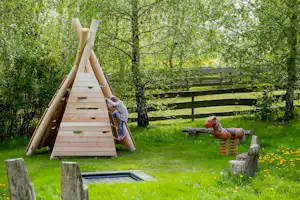 Spielbereich im Grünen: Holz-Klettergerüst, Trampolin und Sitzbank zwischen Bäumen und grüner Wiese.