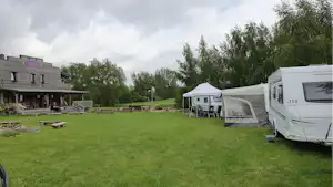 Wohnwagen und Zelt auf einem grünen Campingplatz, daneben ein Gebäude mit Terrasse und Rasenfläche.