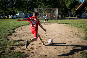 Ein Kind spielt Fußball auf einem Campingplatz-Fußballfeld vor dem Tor, im Hintergrund weitere Kinder und Bäume.