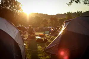 Zelte auf einem Campingplatz bei Sonnenuntergang, im Hintergrund Tische und Stühle zwischen Bäumen.
