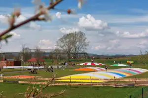 Buntes Springkissen auf dem Campingplatz, daneben Sitzbänke und Spielbereich, im Hintergrund Wiese, Zaun und Bäume.