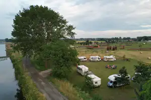 Campingplatz am Fluss mit Wiesen und mehreren Wohnwagen, umgeben von Bäumen und offenen Feldern.