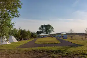 Campingplatz mit Zelt am Rand, gepflastertem Weg und Rasenparzellen, daneben Wohnwagen und Bäume am Feldrand.