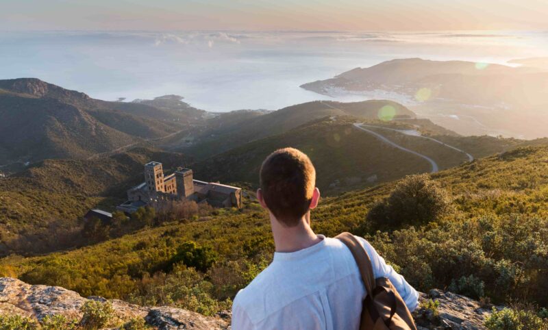 Junger Mann betrachtet das Kloster Sant Pere de Rodes mit dem Mittelmeer im Hintergrund